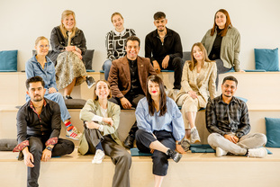 Das Foto zeigt eine Gruppe von Menschen unterschiedlichen Alters, die auf einer Treppe sitzen und in die Kamera gucken.