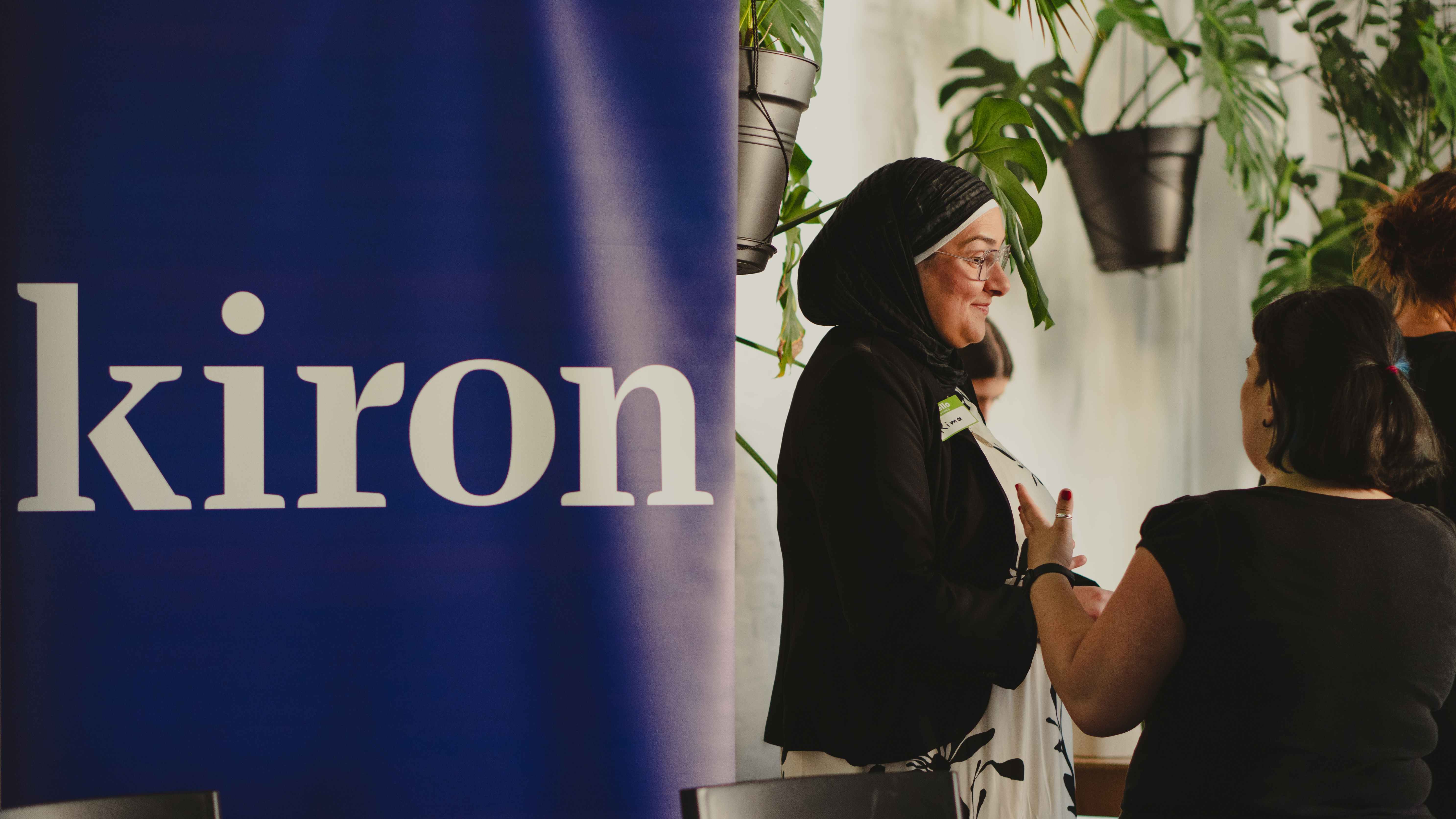 Auf dem Foto ist der Schriftzug der Firma Kiron erkennbar. Zudem sieht man eine Frau mit Kopftuch, die mit anderen Frauen im Gespräch ist.
